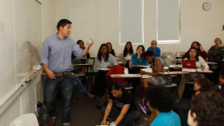 Students sit at desk and watch teacher at white board